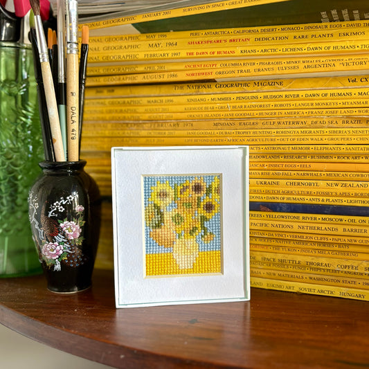 Framed cross-stitch of Van Gogh's 'Sunflowers' on a wooden surface with art supplies.