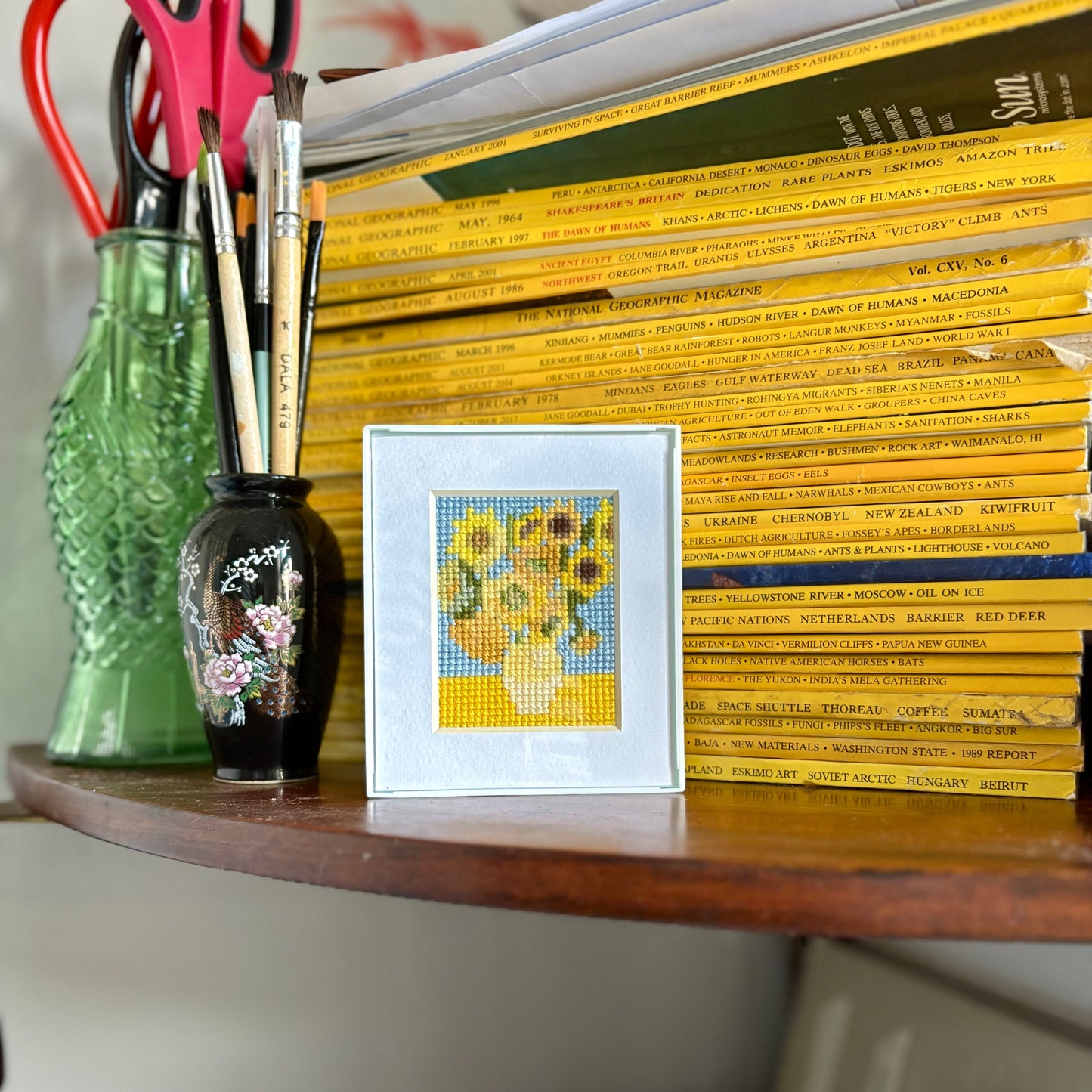 Stack of books on a shelf with a framed cross-stitch picture of sunflowers.