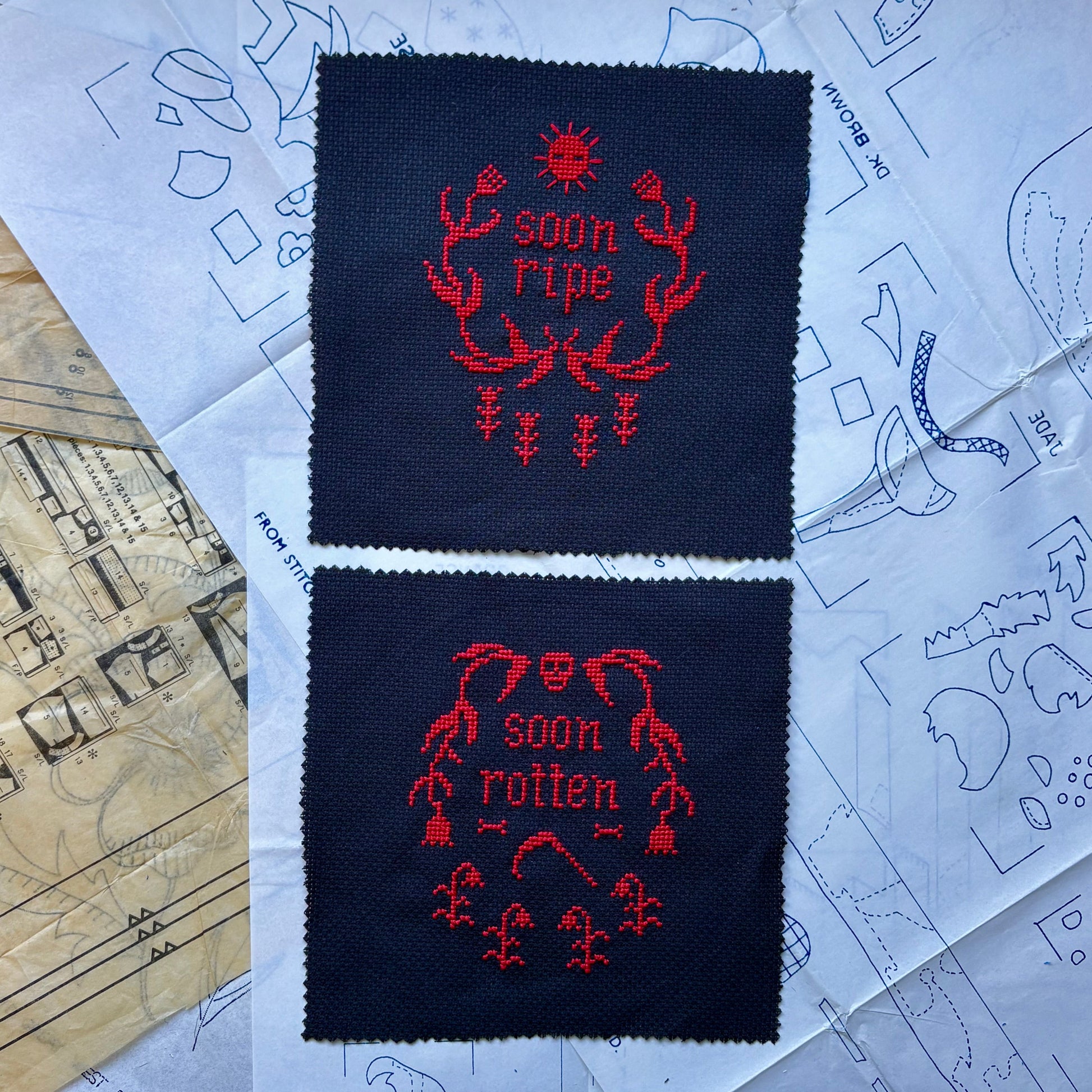 Two embroidered squares with red text on navy fabric, placed on a paper with design sketches.
