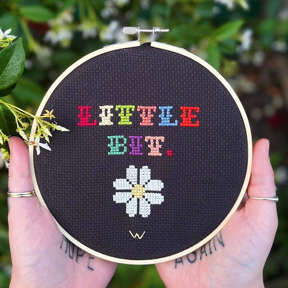 Embroidery hoop with colorful text and a white flower design held by hands against a floral background