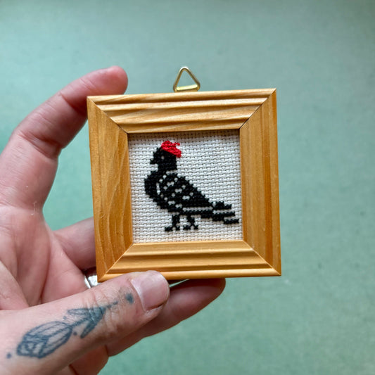 Hand holding a small wooden frame with a cross-stitched pigeon ornament against a green background