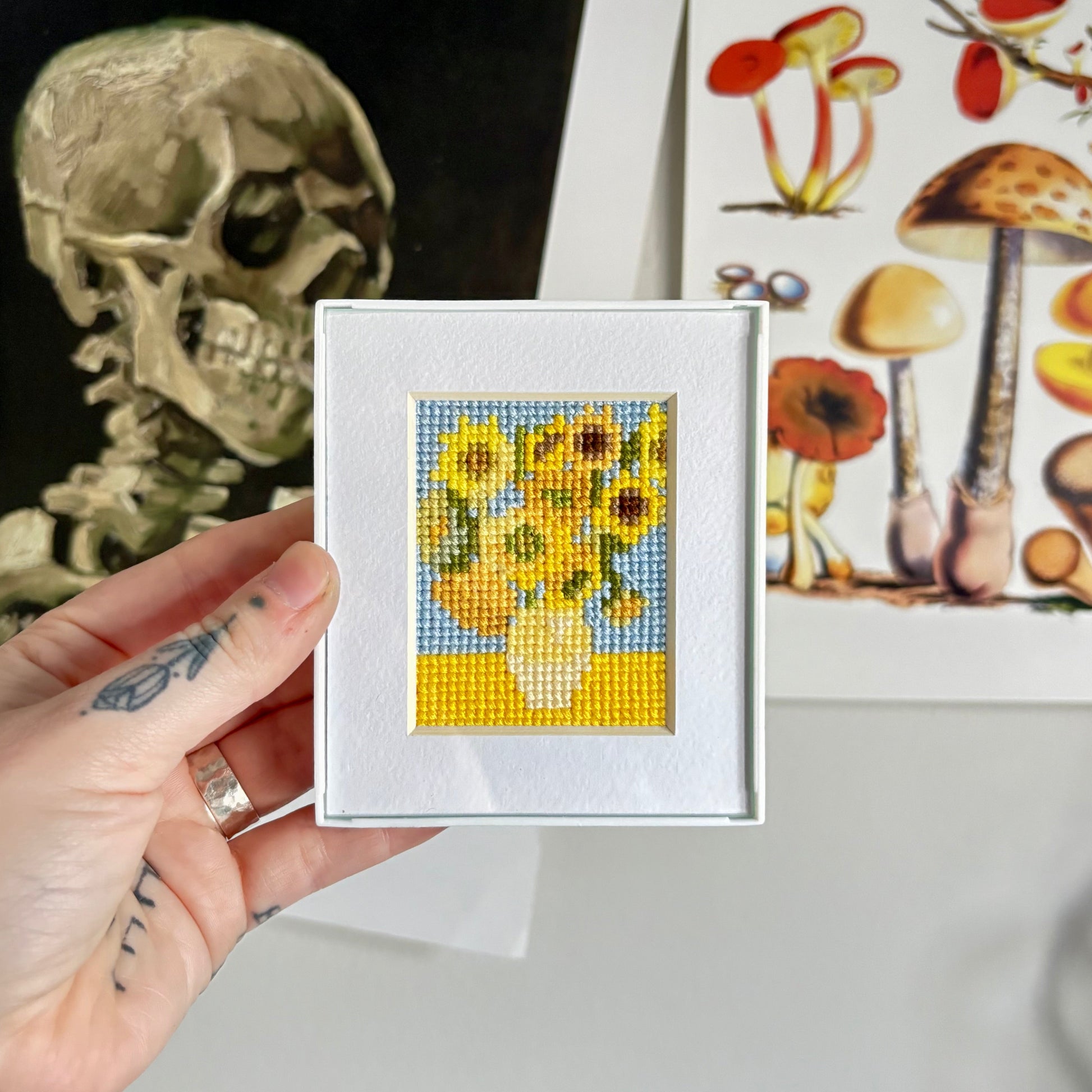 Hand holding a small cross-stitched piece of art with sunflowers, with a skull and mushroom illustration in the background.
