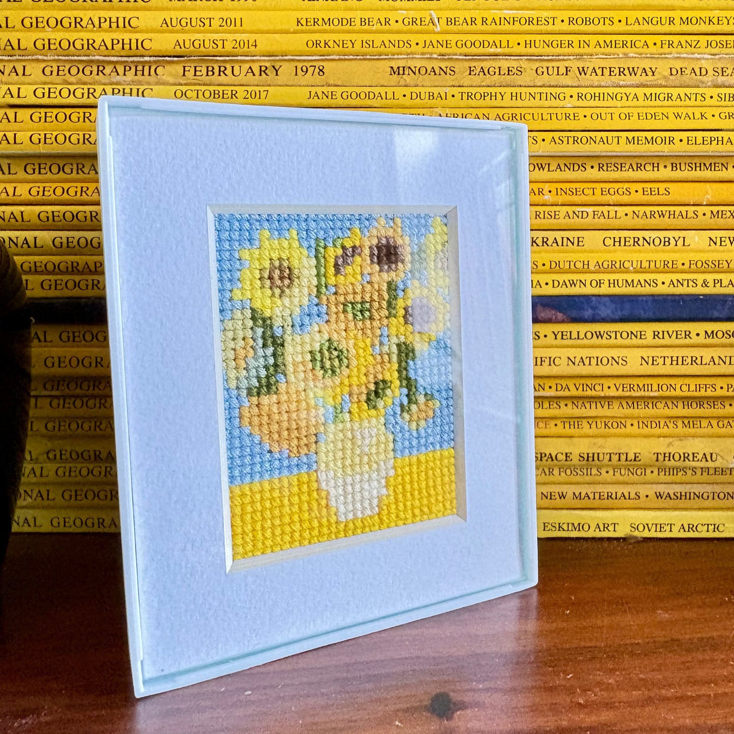 Cross-stitched sunflower design on a card with National Geographic magazines in the background