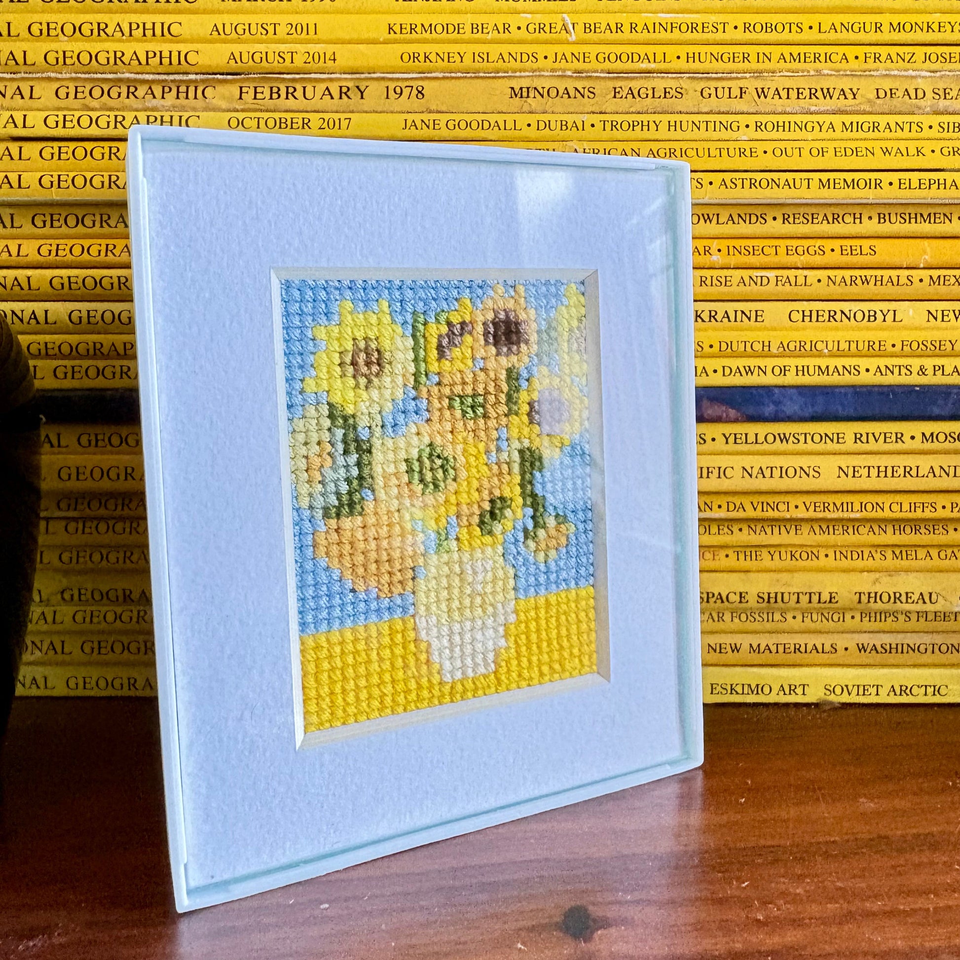 Cross-stitched sunflower design on a card with National Geographic magazines in the background