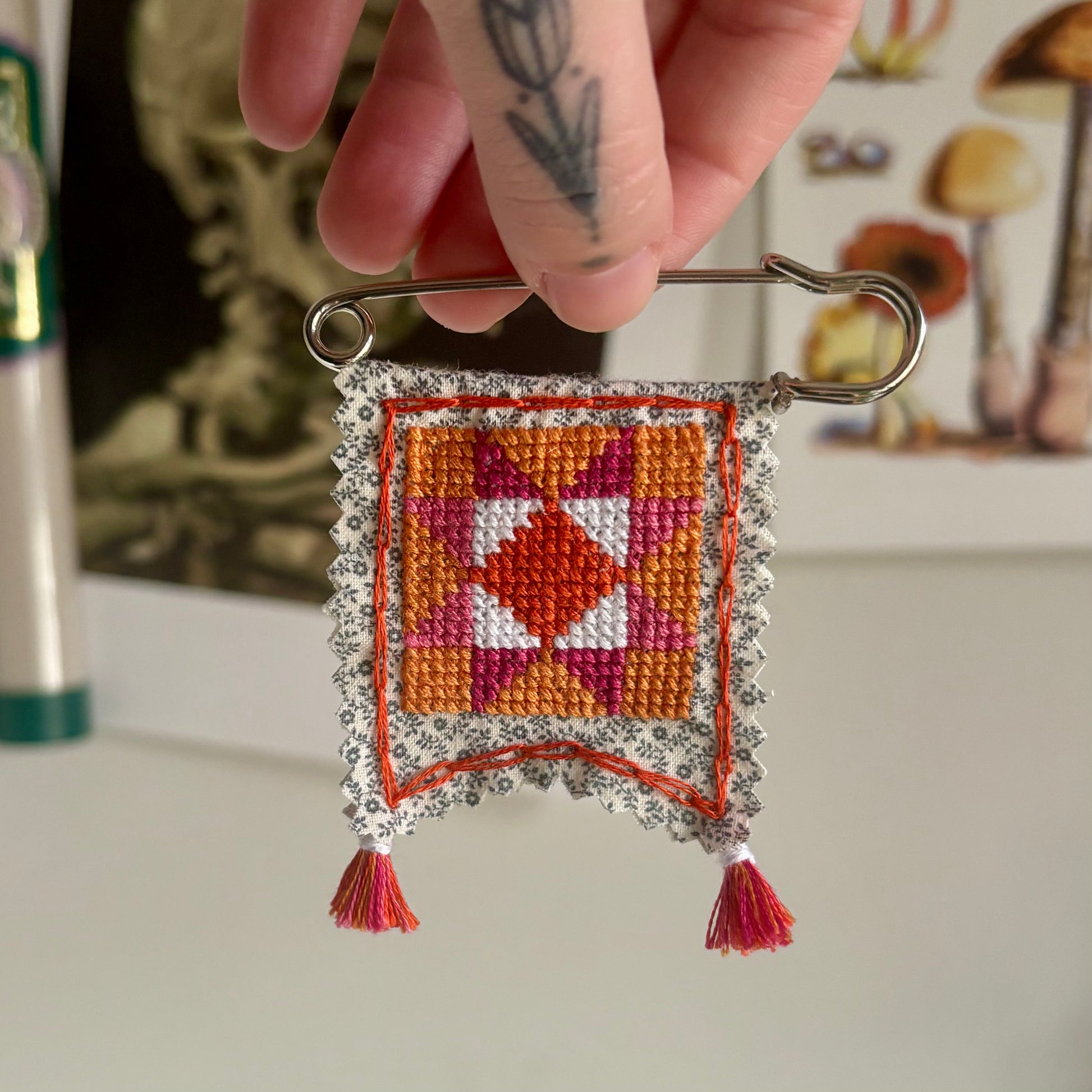 Handmade cross-stitched decorative item on a safety pin against a blurred background