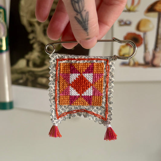 Handmade cross-stitched decorative item on a safety pin against a blurred background