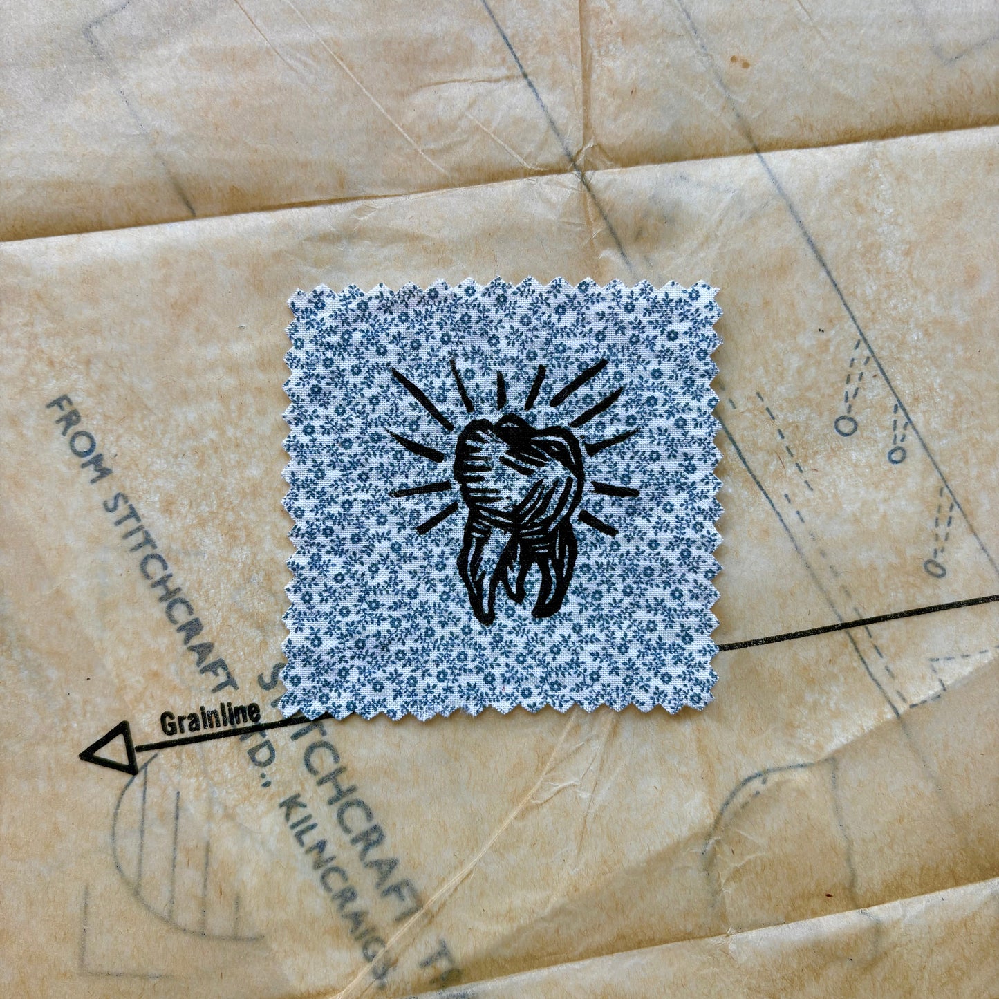 Square patch with black design on a floral pattern, placed on a textured surface with text.