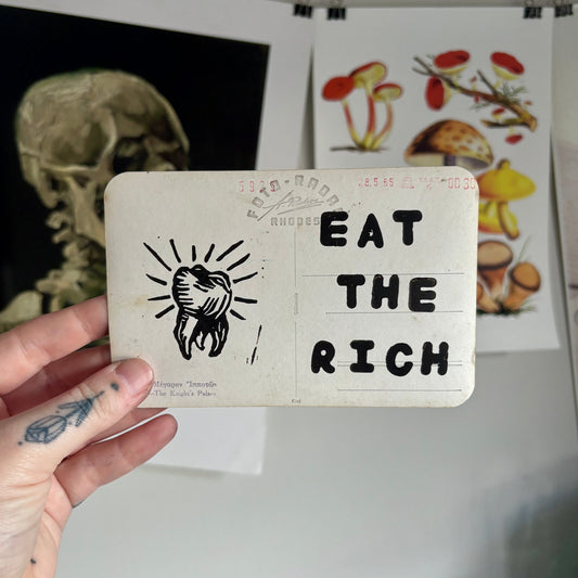Hand holding a card with 'Eat the Rich' text and a skull illustration, with a blurred background of artwork.